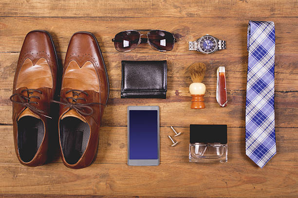 Variety of men's accessories organized in knolling arrangement.  Items include: dress shoes, tie, sunglasses, wallet, watch, shaving brush, cologne, and smart phone.  The items all lie on a wooden desk or table.   Men's personal accessories, clothing themes.  Father's Day.  Fashion.  Business.  Retail.